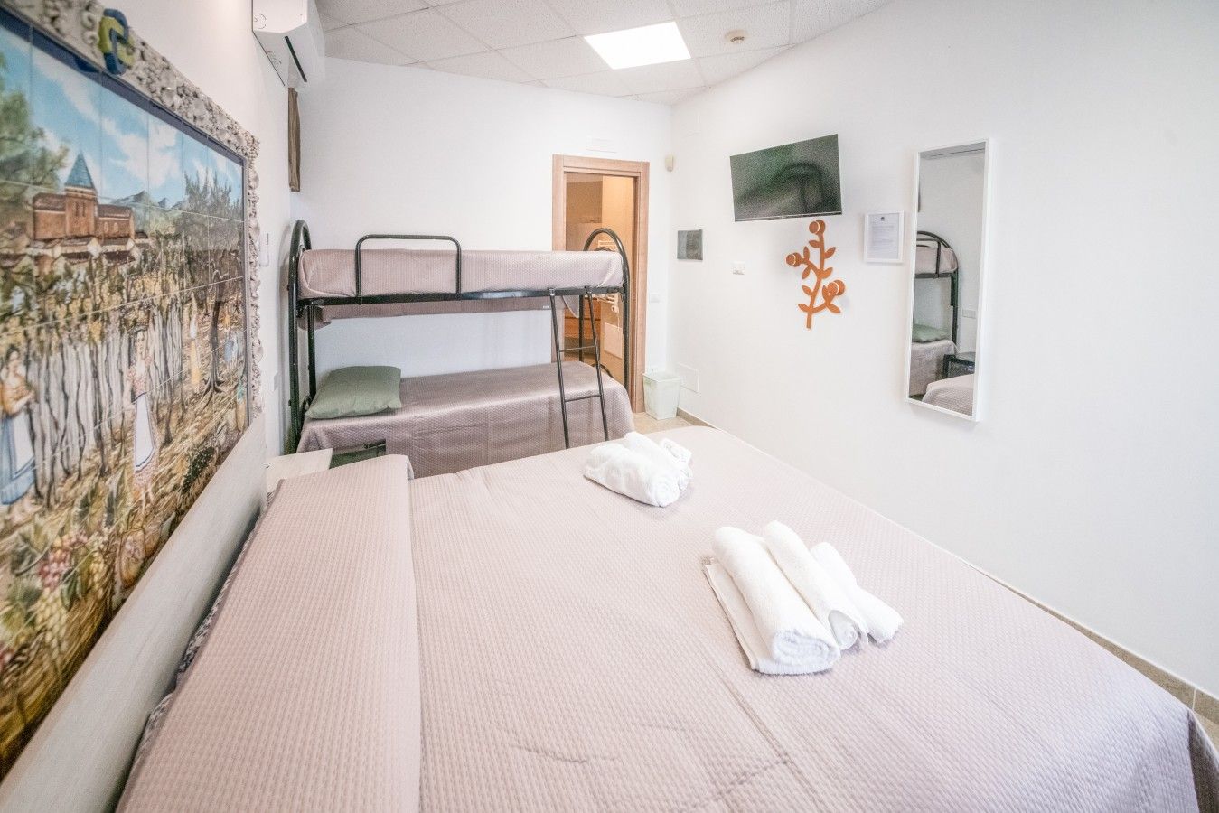 Hotellihuone, jossa on kerrossängyt, sänky, jossa on rullalle taiteltuja pyyhkeitä, taulu, televisio ja peili.