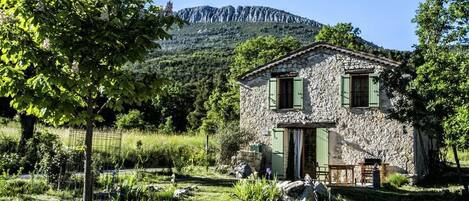 Ein Steinhaus mit Tür und Fenstern, umgeben von Grün und einem Berg im Hintergrund.