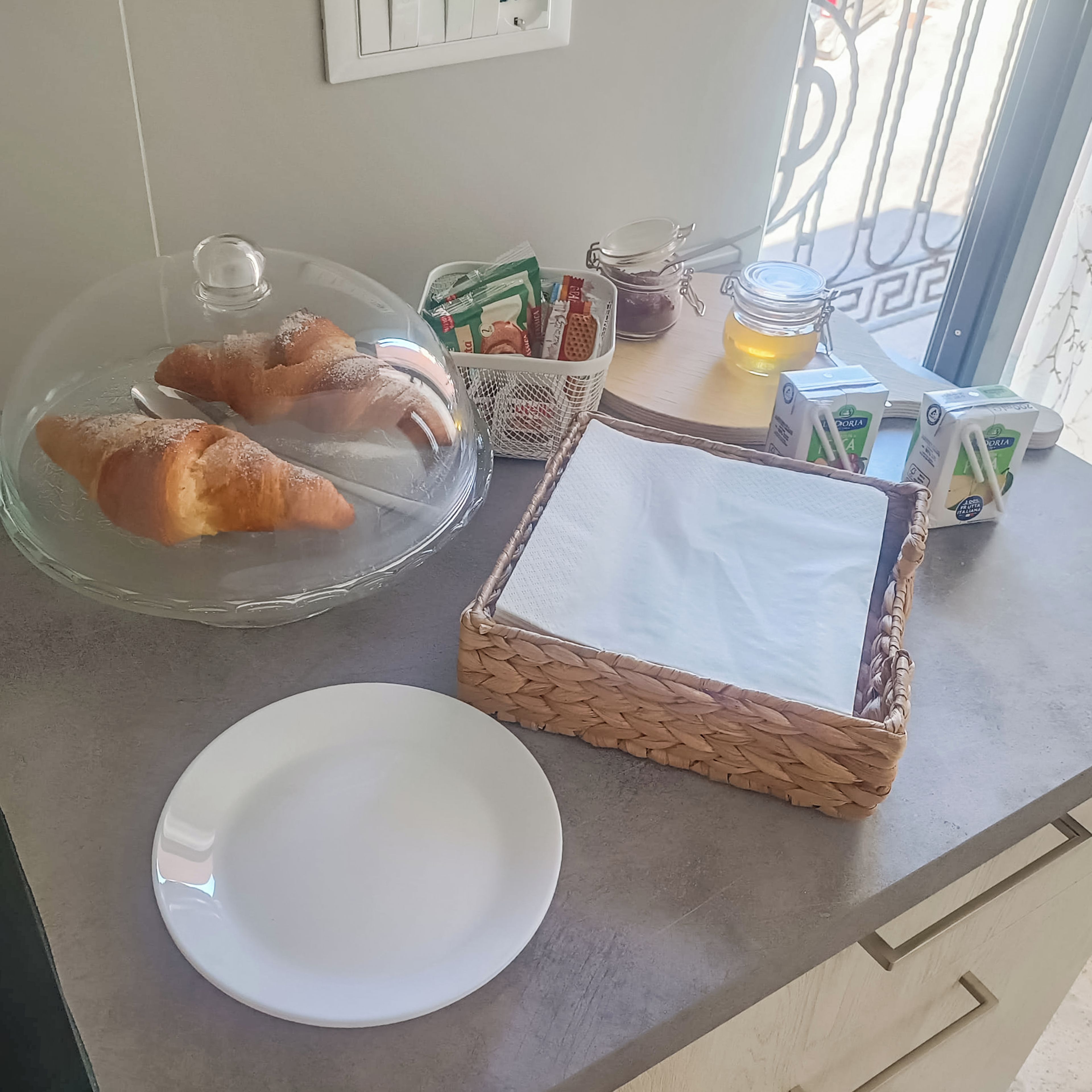Ontbijt met croissants, een glazen houder, een mandje, een pot en pakken melk op een aanrecht.