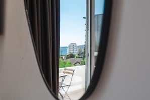 A view through a window with a chair and table on a balcony, overlooking a building and a body of water. - Andrea Boutique Hotel (Sarande)