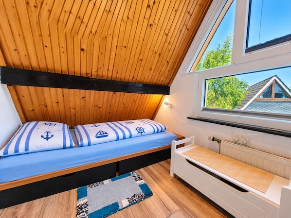 Habitació petita i acollidora amb sostre inclinat, un llit individual amb roba de llit de temàtica marina, una finestra amb vista a una casa i un radiador.