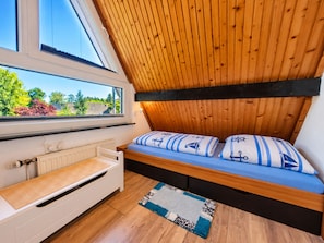 Een kamer met een schuine kap, een bed met nautisch beddengoed, een raam met uitzicht op groen en een radiator.