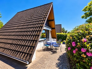 Een huis met een schuin dak, een terras met buitenmeubilair en een tuin met bloeiende planten.
