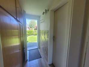 Un passadís amb una porta de fusta, una porta de vidre que dona a l'exterior i una paret enrajolada amb un llum.