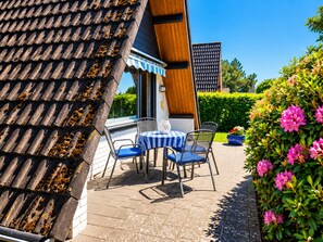Een terras met tafel en stoelen, een pannendak en een haag met roze bloemen.