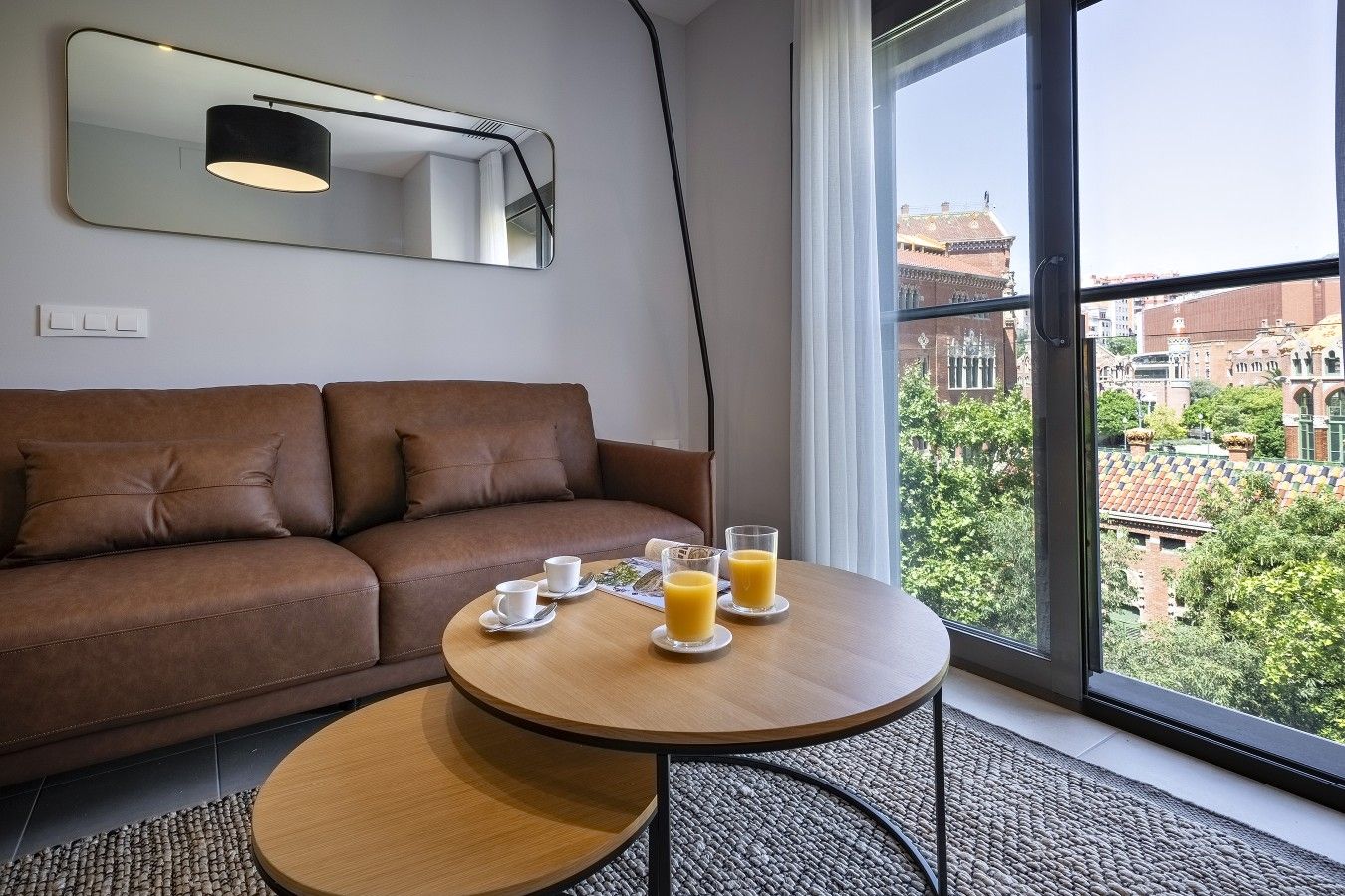 Ein Zimmer mit einem braunen Sofa, einem runden Holztisch mit zwei Tassen Kaffee und zwei Gläsern Orangensaft, einem Spiegel über dem Sofa und einem Fenster mit Blick auf Gebäude und Bäume.