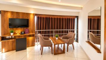 A modern hotel room with a wooden reception desk, a flat-screen TV, a dining table with chairs, and a large window with curtains.