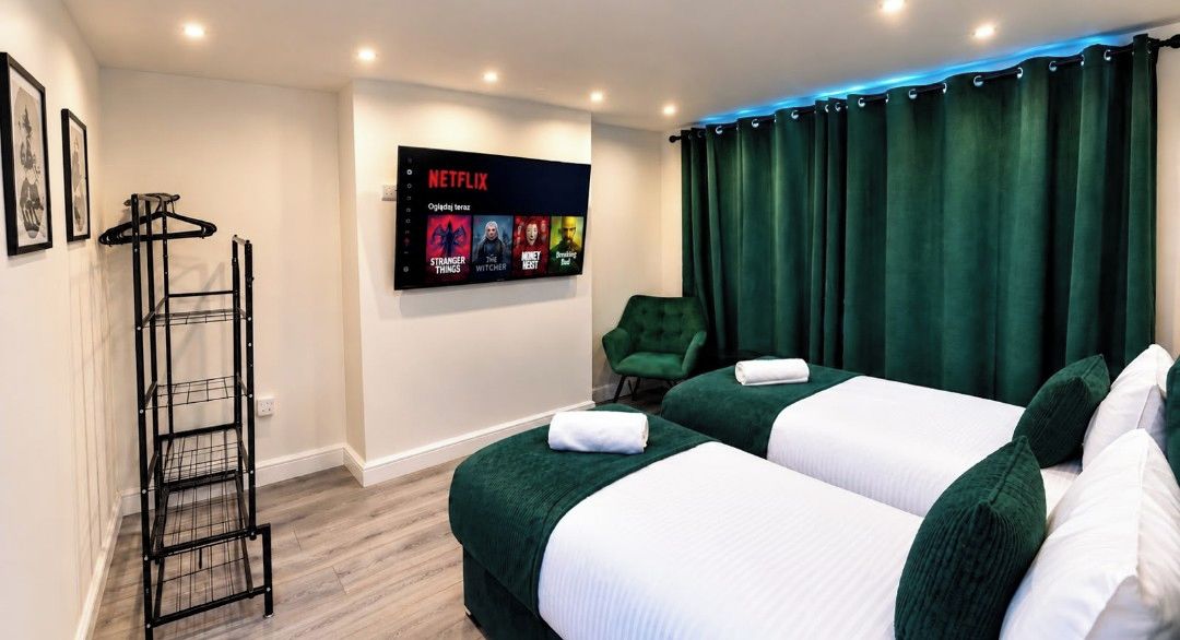 Une chambre d’hôtel avec deux lits, un fauteuil vert, un téléviseur diffusant du contenu Netflix et une étagère en métal noir.