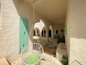 A hanging chair with a green cushion in a sunlit courtyard with stone flooring, a turquoise door, and a white wall. - Dar Oueghlani (El May)