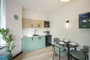 Uma cozinha moderna com pia, micro-ondas e máquina de café, uma mesa de jantar com cadeiras e uma TV fixada na parede exibindo o aplicativo VIDAA.