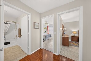 Un couloir avec une salle de bain sur la gauche, une chambre avec un lit et une fenêtre sur la droite, et une table de chevet avec une lampe.