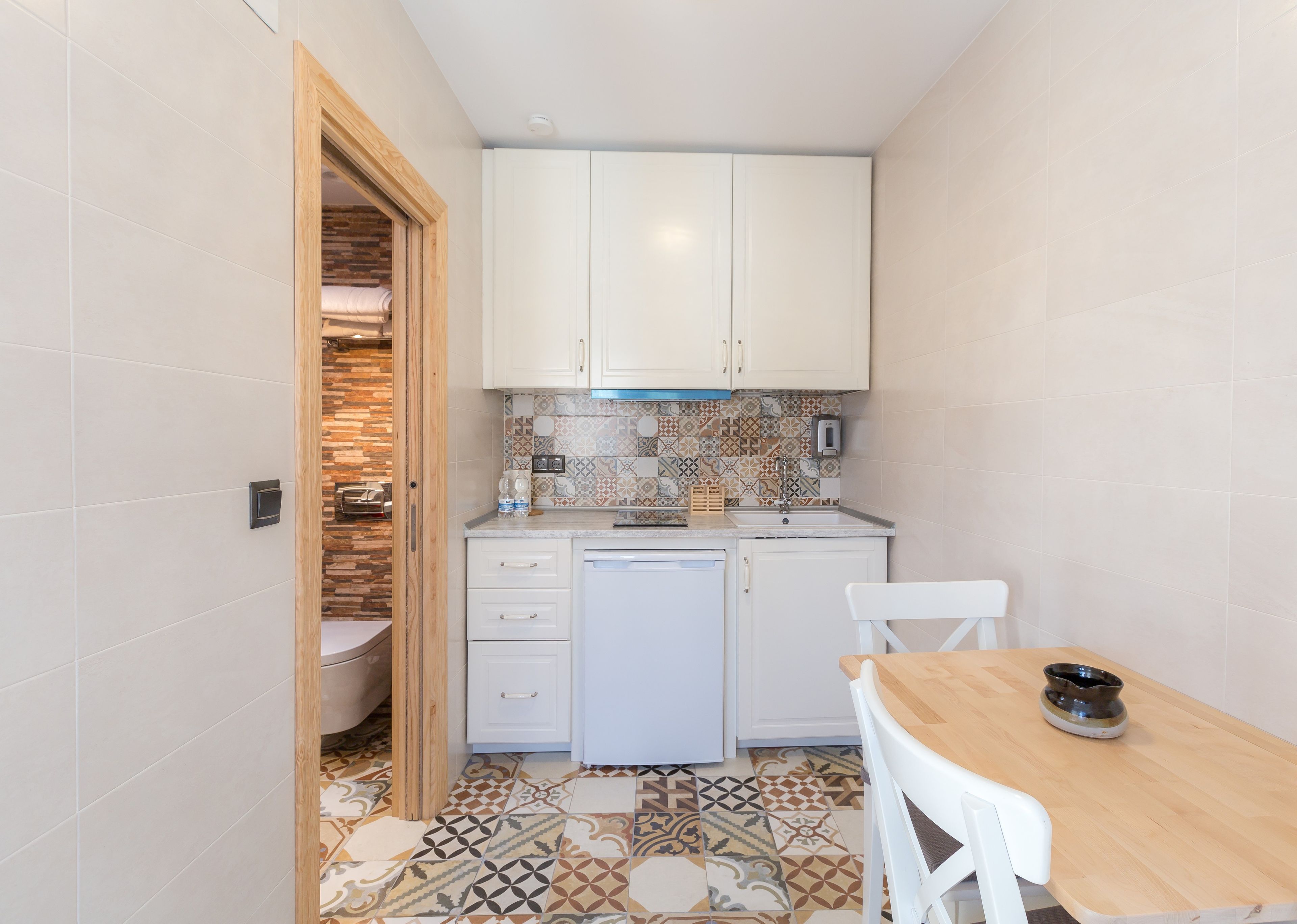 Una cocina compacta con gabinetes blancos, un salpicadero estampado y suelo de baldosas.