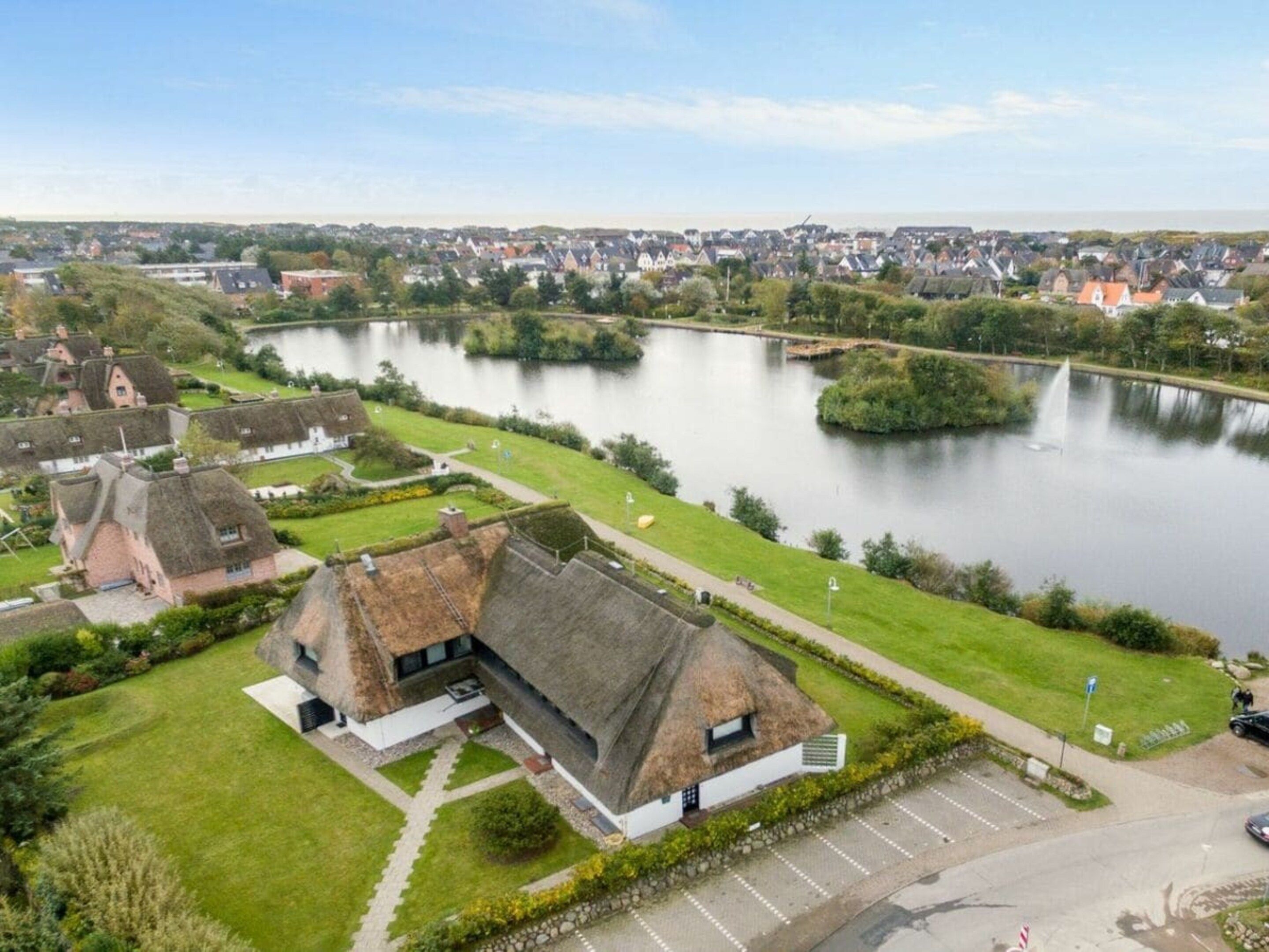 Une propriété située au bord de la rivière, avec un bâtiment à toiture de chaume, un espace de stationnement et une vue sur une ville de l’autre côté de l’eau.