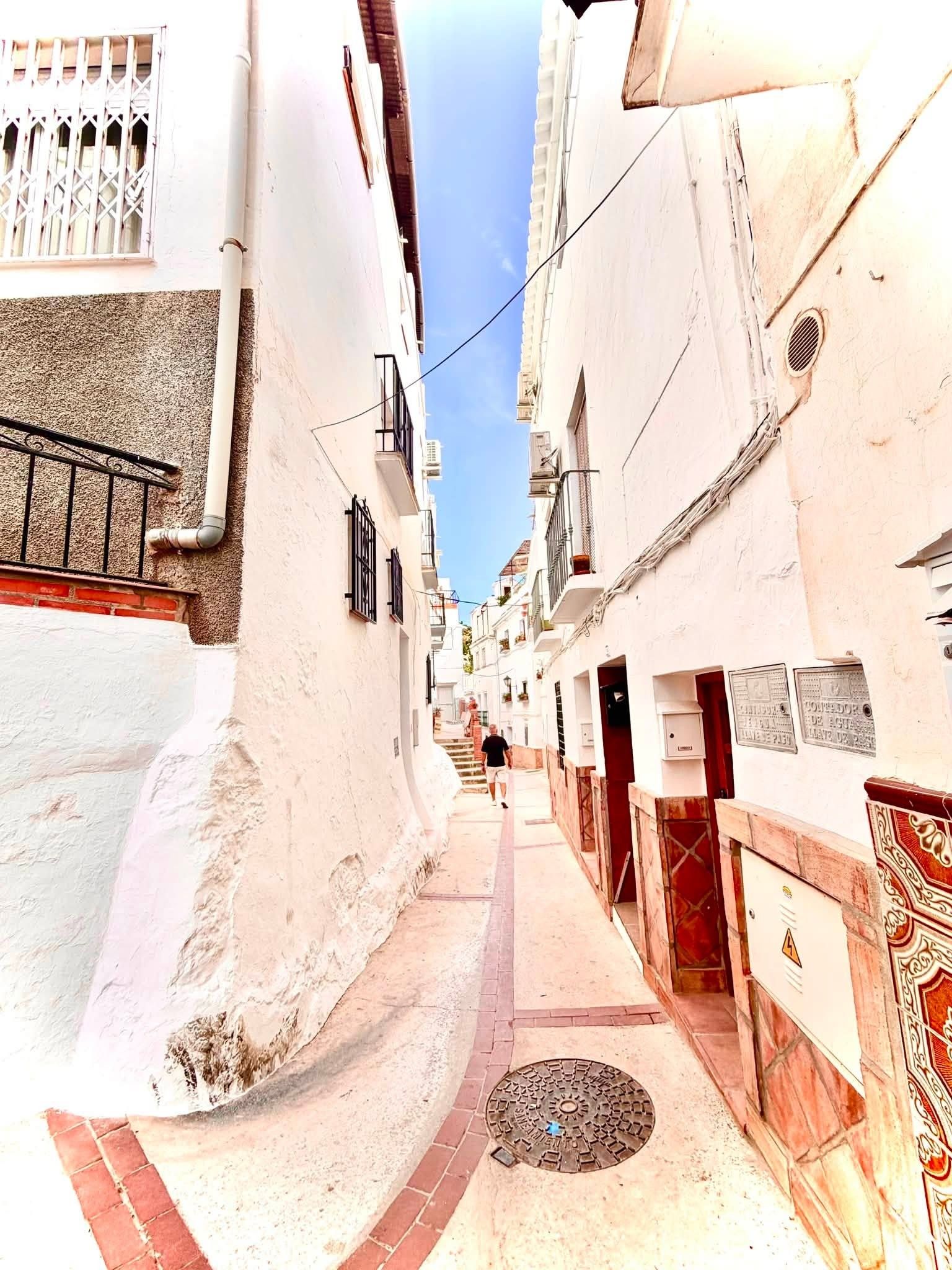 Una calle estrecha con edificios blancos, una tapa de registro y una valla metálica.