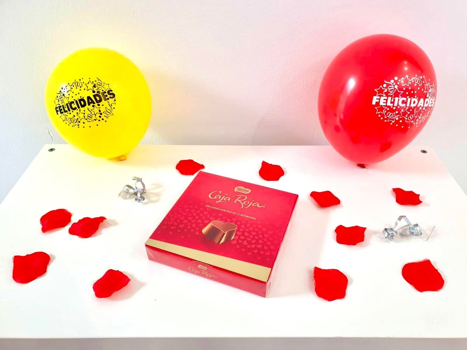 Ein Tisch mit einer roten Schachtel mit dem Aufdruck 'Caja Roja' und zwei Ballons mit der Aufschrift 'FELICIDADES'.