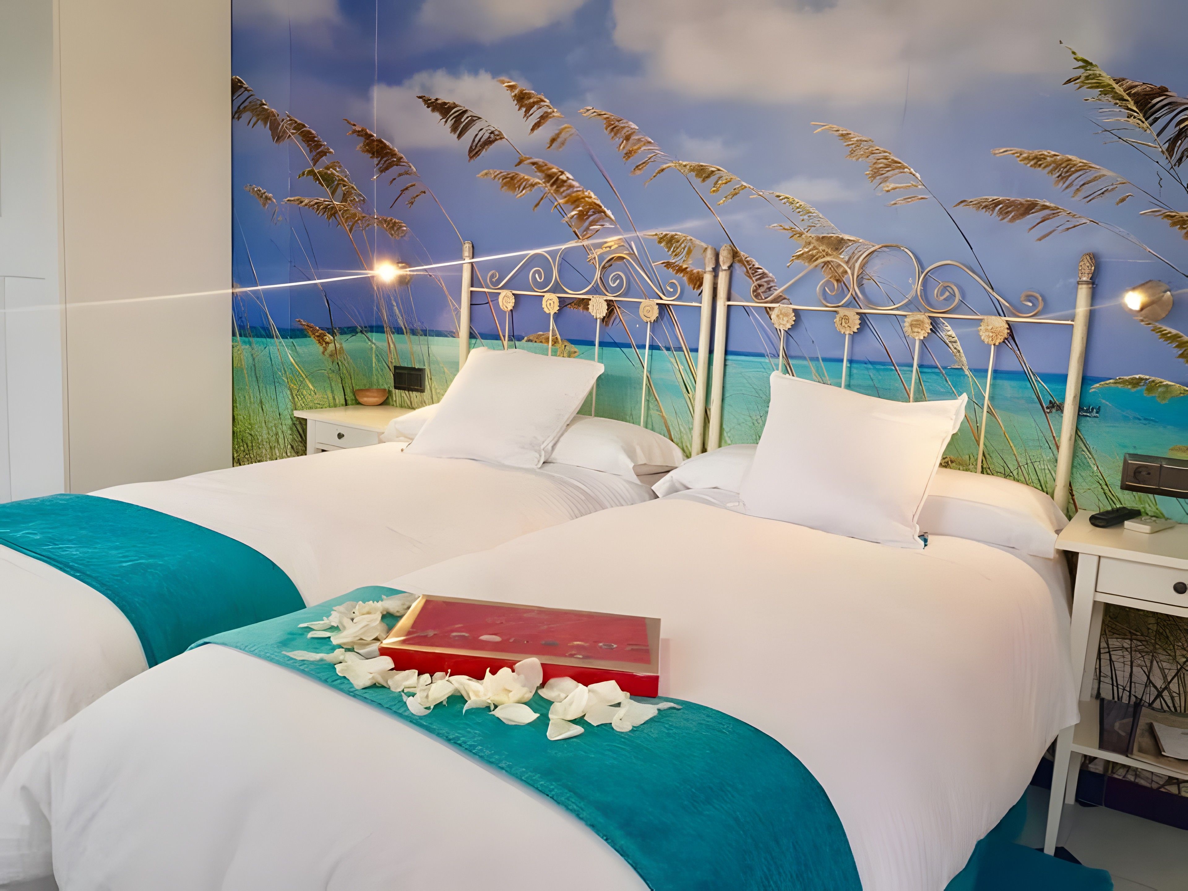 Una cama doble con ropa blanca y un detalle verde azulado, un libro rojo y pétalos blancos esparcidos sobre la cama.