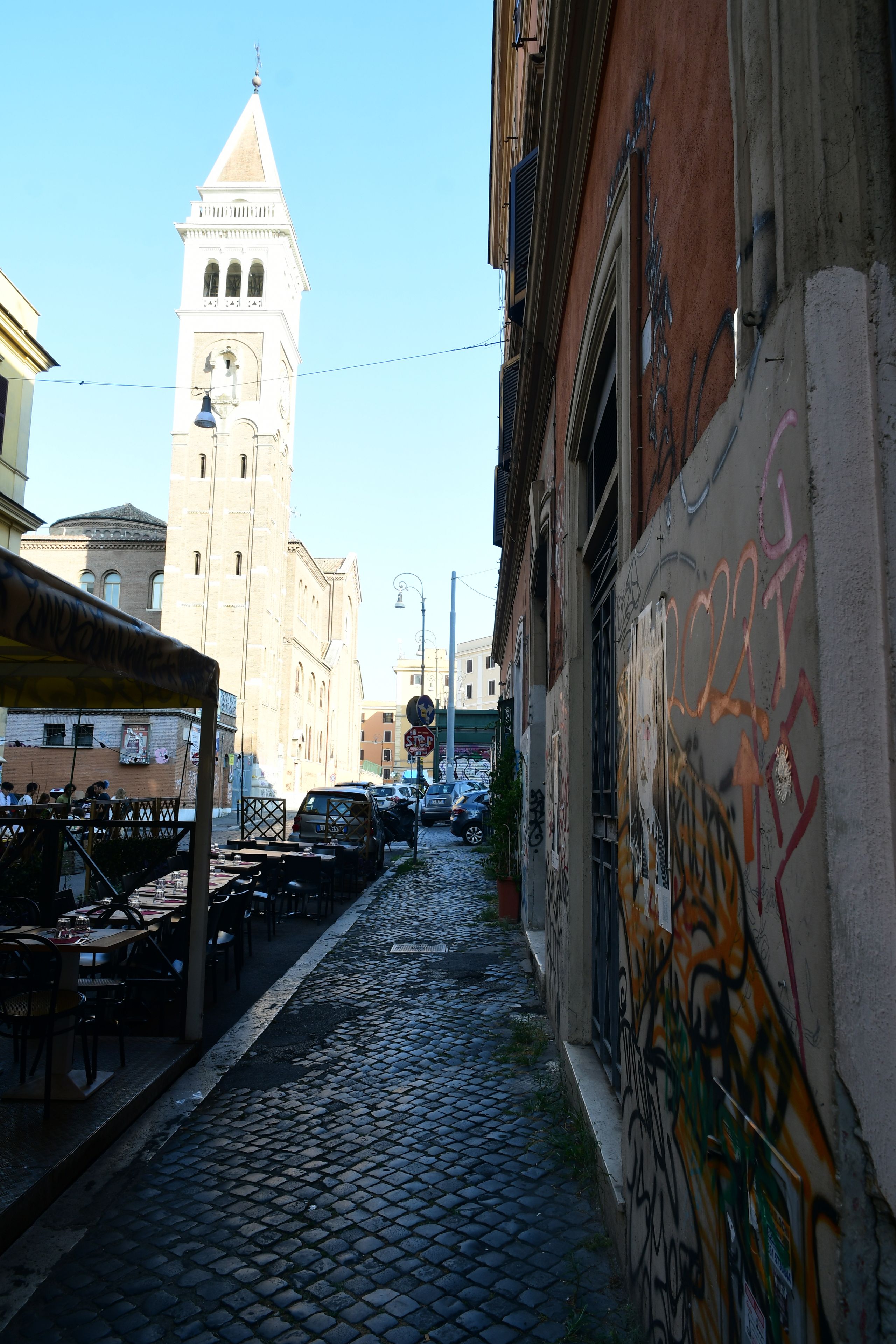 Una estrecha calle adoquinada con una torre al fondo, un edificio con grafitis a la derecha y un área de estar al aire libre techada a la izquierda.
