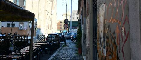 Una estrecha calle adoquinada con una torre al fondo, un edificio con grafitis a la derecha y un área de estar al aire libre techada a la izquierda.