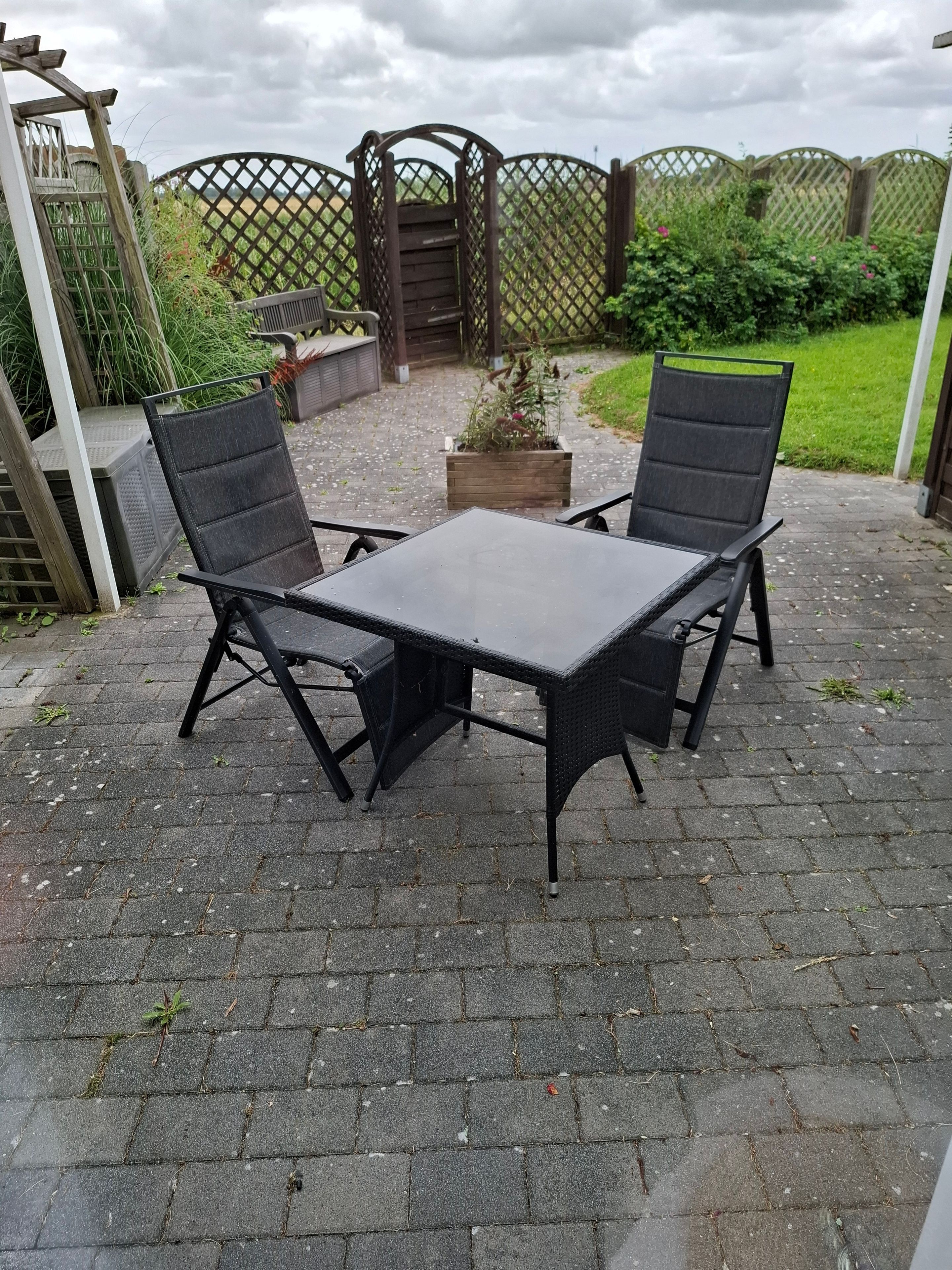 Um pátio externo com uma mesa e duas cadeiras, cercado por um muro de madeira e vegetação.