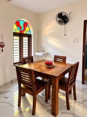 Un comedor con una mesa y sillas de madera, una ventana con un diseño de vitral, un ventilador montado en la pared y un lavamanos pequeño blanco.