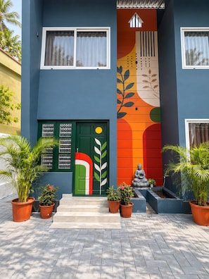 A building with a colorful mural, a green door with a circular window, and potted plants in front. - The Village House (Varkala)