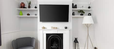 A living room with a fireplace, a flat-screen TV mounted above it, a grey armchair, a dark sofa, and a floor lamp.