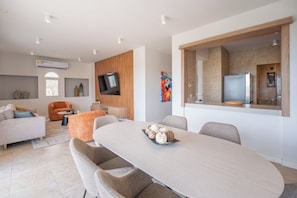 Una sala de estar moderna con comedor, un televisor de pantalla plana montado en la pared y una cocina visible a través de un vano abierto.