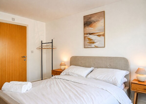 A bedroom with a bed, a wooden door, a wall-mounted artwork, and a nightstand with a lamp. - Stylish 2-Bed, 2-Bath Flat with Balcony & Parking (Warrington)