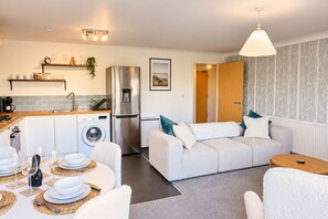 A compact living space with a kitchenette, a dining area set for four, and a sofa with decorative pillows. - Stylish 2-Bed, 2-Bath Flat with Balcony & Parking (Warrington)