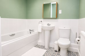 Un baño con bañera, lavabo e inodoro, con sanitarios blancos y baldosas de suelo estampadas.