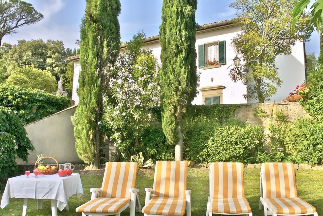 A patio with four striped lounge chairs, a table with a basket of fruit, and a white building with green shutters in the background.