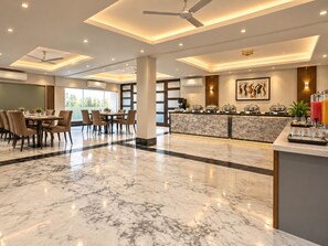 Un vestíbulo de hotel con piso de mármol, una zona de comedor con sillas y una mesa, una recepción con mostrador y un ventilador de techo.