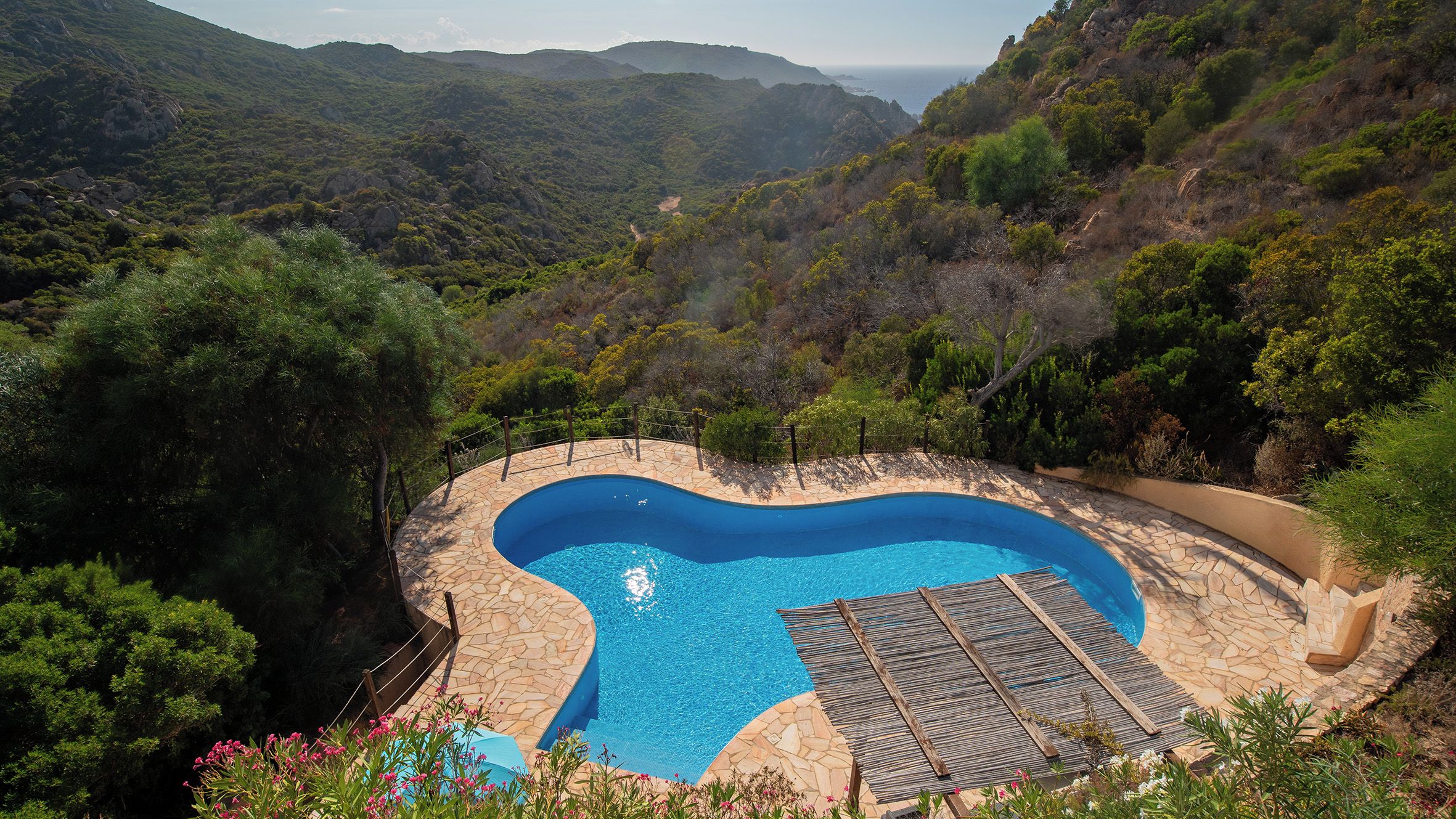 Une piscine dotée d’une plage en pierre, entourée d’une végétation luxuriante et de collines.
