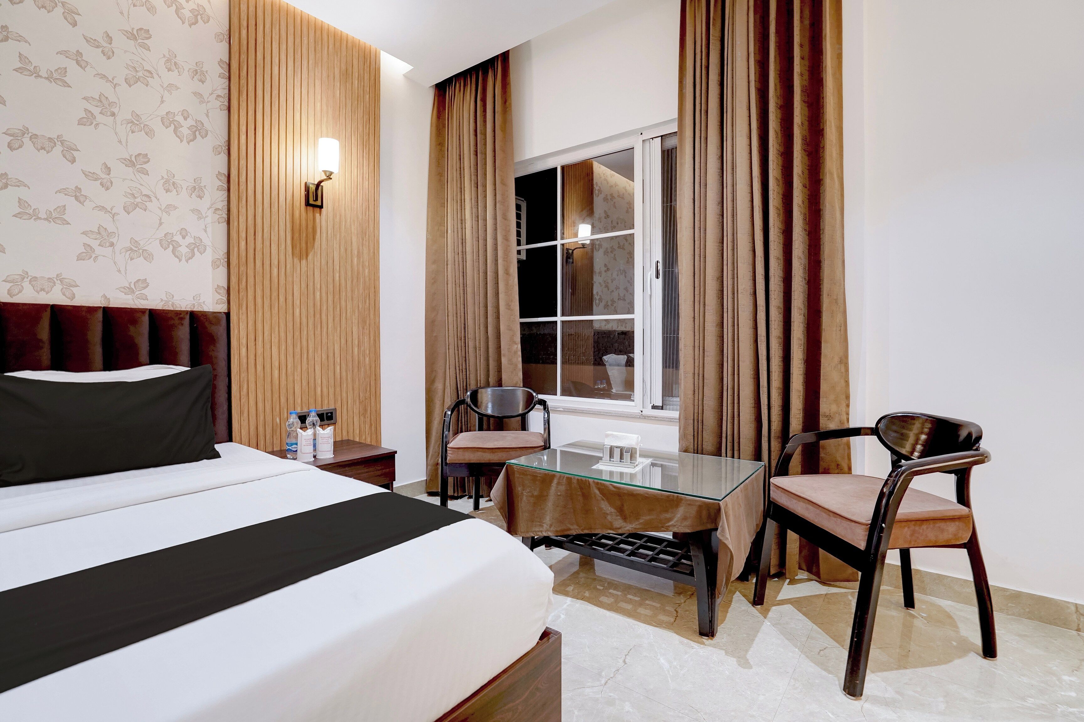 A hotel room with a bed, a desk, two chairs, a window, and a wall with vertical wooden panels.