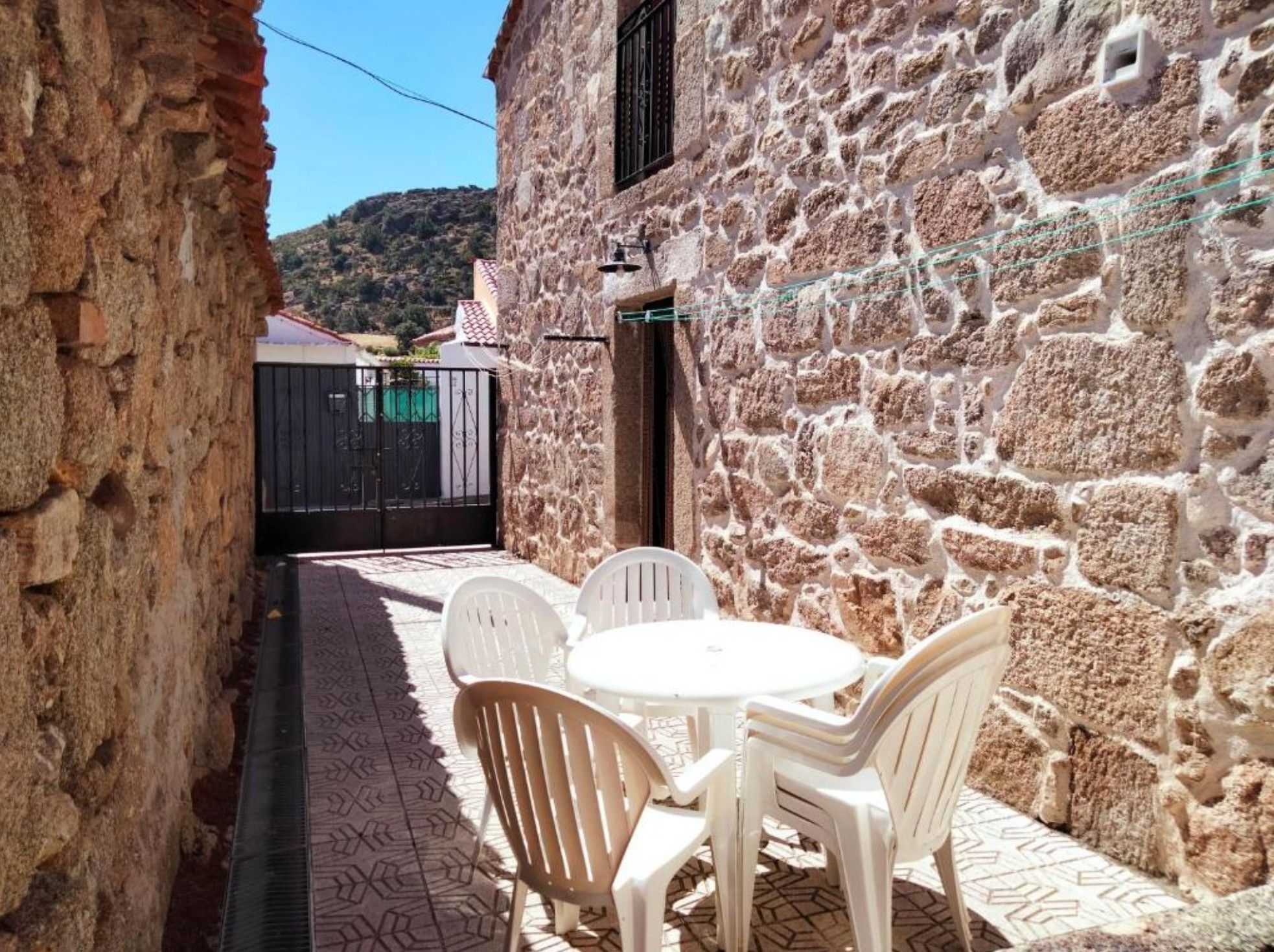 A narrow alleyway with a stone wall on one side and a gate leading to a building. There is a small round table with chairs in the foreground.