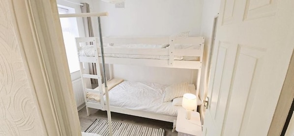 A bunk bed room with a white door, a bedside table with a lamp, and a striped rug on the floor. - The Charlton Suite (Harrow)