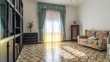 A room with patterned floor tiles, a sofa with floral upholstery, a cabinet with glass doors, and a window with sheer curtains.