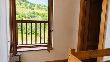 Una habitación con piso de madera, una ventana con cortinas abiertas, vista a un paisaje verde y una puerta.