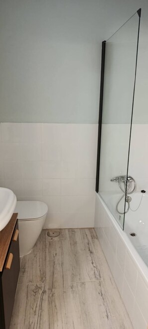 Een badkamer met een toilet, een houten wastafelmeubel en een badkuip met glazen douchewand.