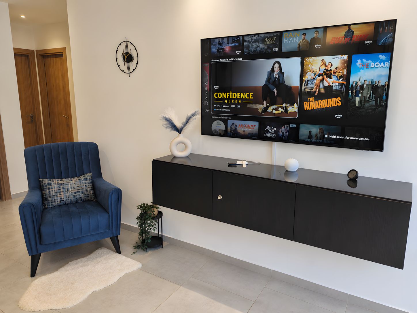 Un salon moderne avec un téléviseur à écran plat fixé sur une console noire, un fauteuil bleu et une horloge murale décorative.