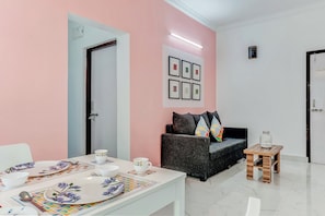 A room with a dining table set with plates, cups, and cutlery. There is a sofa with cushions and a small wooden table with a glass jar. - Hotel O Dazzling Studio (Pune)