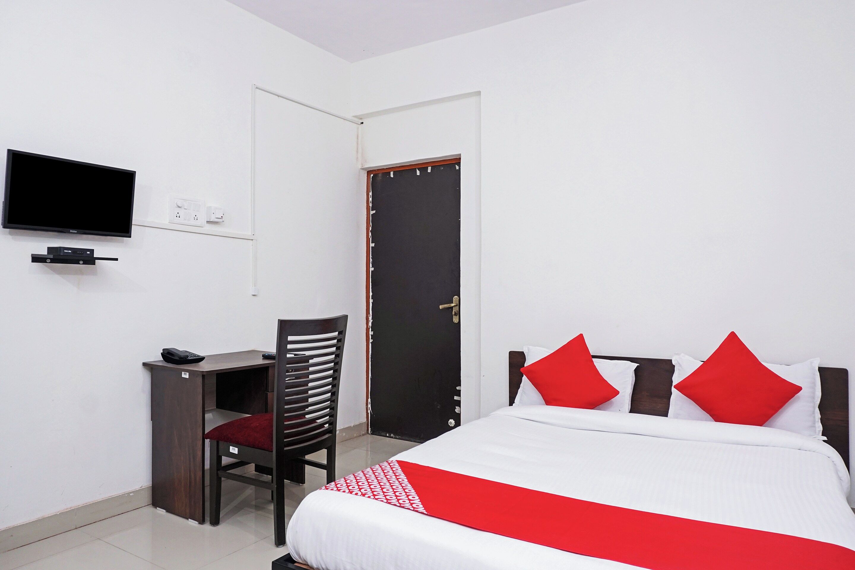 Quarto de hotel com cama, escrivaninha, cadeira, televisão e porta.