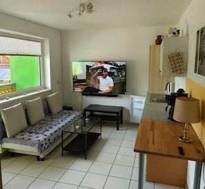 Een woonkamer met een bank, een salontafel, een aan de muur bevestigde televisie, een keukenhoek met een spoelbak en een raam met zonwering.