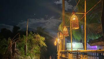 A nighttime scene with a pathway flanked by bamboo railings and hanging lanterns, leading to a building with a green-lit facade.