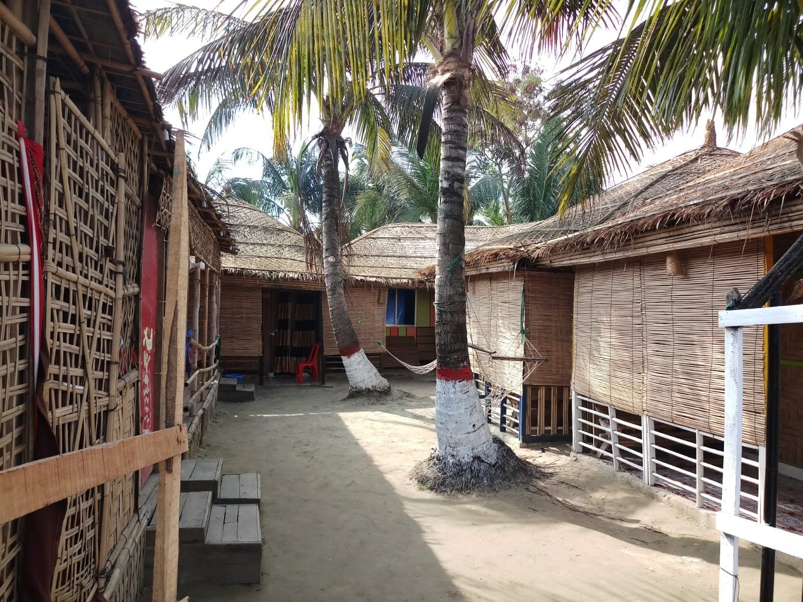 A traditional thatched-roof structure with a central open doorway, surrounded by palm trees and a sandy ground.