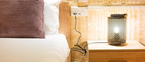 A bed with a white pillow and a brown pillow, a wooden nightstand with a lamp, and a wall socket.