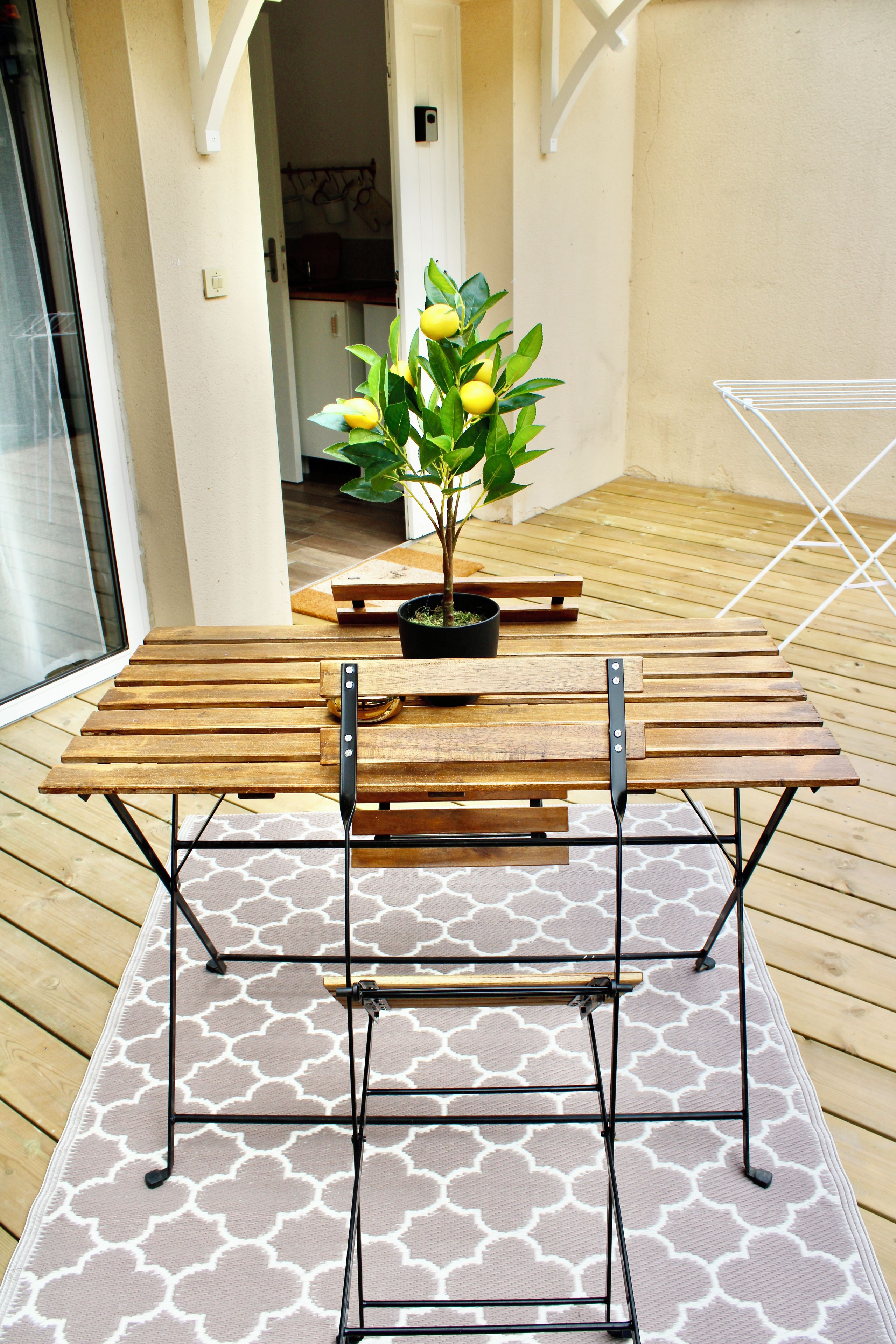 An outdoor wooden table with metal legs, a potted plant with yellow flowers, and a patterned rug on a wooden deck.