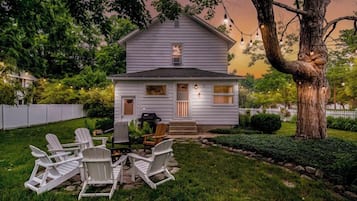 A white house with a porch, outdoor seating, and a large tree.