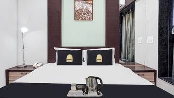 A hotel room with a bed, two pillows with 'OAK' logo, a bedside lamp, a telephone, a tray with a coffee pot and cups, and a painting on the wall.