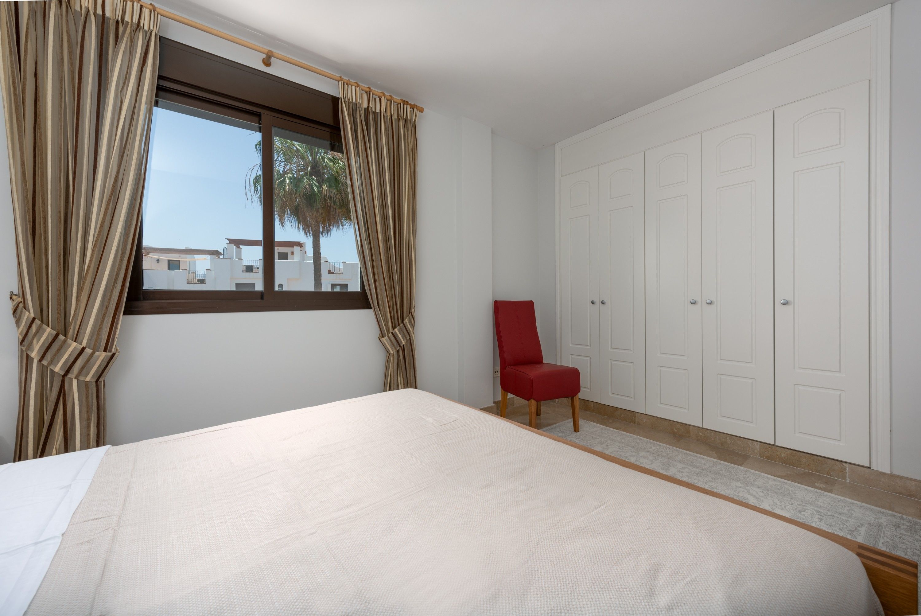 Een slaapkamer met een bed, een rode stoel, een kledingkast en een raam met gordijnen.